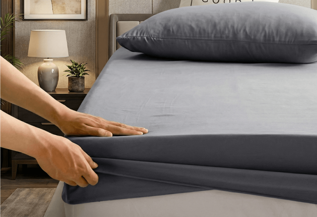 Person adjusting gray fitted sheet on a bed in a bedroom setting.